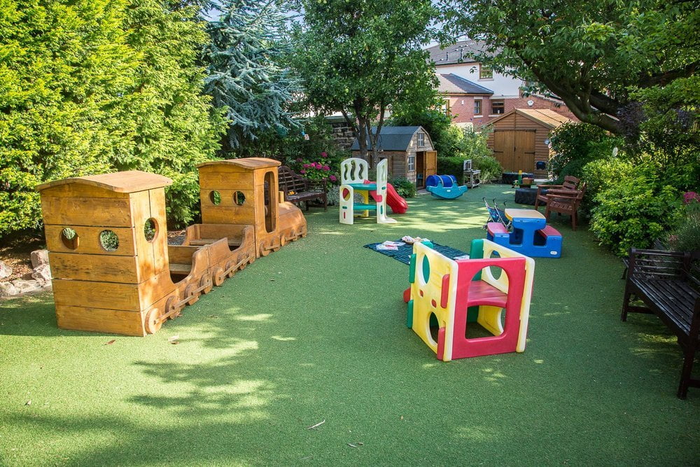 A nursery garden in the sun: a wooden play train, a small slide and play equipment on artificial grass, with trees behind.